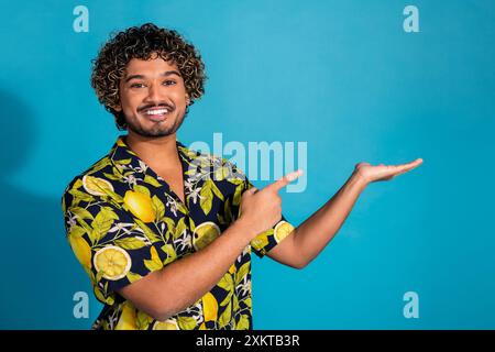 Foto eines guten Mannes mit gewelltem Haar gekleidetem Print-Hemd, das auf Objekt auf Arm leeren Raum lokalisiert auf blauem Hintergrund zeigt Stockfoto