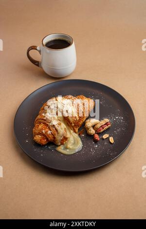 Croissant mit süßer Füllung und Mandeln mit Puderzucker liegt auf schwarzer Platte. Lebensmittel Foto Kanne Kaffee auf papierbraunem Hintergrund, Draufsicht Stockfoto