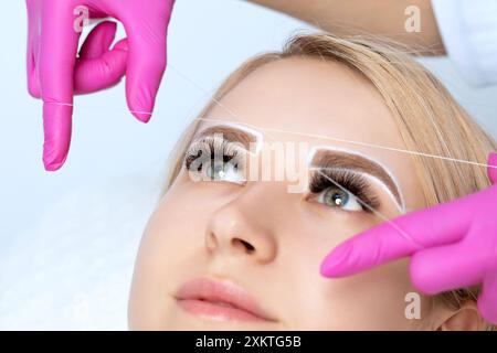 Der Schminkkünstler zupft Augenbrauen mit einer Fadennaht. Frau mit Permanent Make-up Tattoo auf den Augenbrauen. Professionelles Make-up und Kosmetik Stockfoto