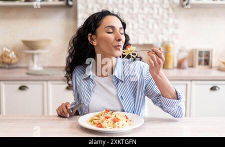 Junge lateinische Frau, die köstliche Pasta isst, leckeres hausgemachtes Mittagessen mit geschlossenen Augen genießt, während sie am Tisch in hellem Küchenraum sitzt, freier Platz Stockfoto