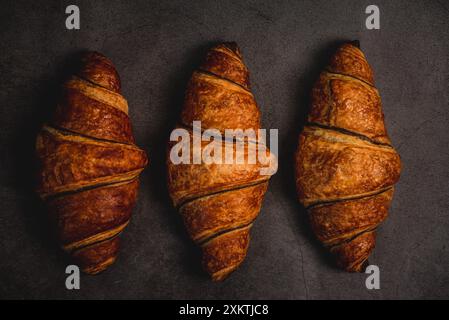 Drei frisch gebackene Croissants sitzen auf einer dunkelgrauen Arbeitsplatte, bereit zum Verzehr. Ihre goldbraune Kruste glitzert unter dem weichen Licht und lädt ein Stockfoto