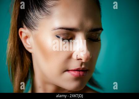 Eine schöne junge Frau mit einem ruhigen Ausdruck und geschlossenen Augen wird vor einem leuchtenden blaugrünen Hintergrund gefangen gehalten. Ihr Haar ist mit einem schlanken Pferdeschwanz gestylt Stockfoto