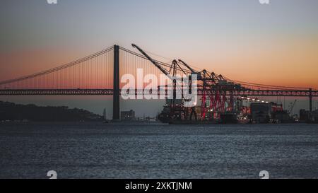 Ein Panoramablick auf einen Schiffshafen mit Kränen und einem Frachtschiff vor dem Hintergrund einer Brücke bei Sonnenuntergang. Der Himmel mit leuchtenden Farben Stockfoto