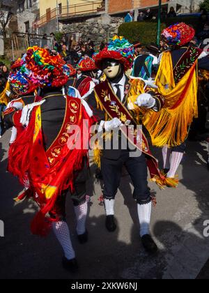 Balari tanzt während des Karnevals in den Straßen von Bagolino, trägt die traditionelle Tracht mit weißen gestrickten Socken, schwarzem Kleid, farbenfrohem Schal über dem Rücken, Gesicht mit elfenbeinfarbener und schwarzer Maske und Kopf unter einem Filzhut mit rotem Band, mit goldenem Schmuck und bunten Bändern, die eine Schleife bilden Stockfoto