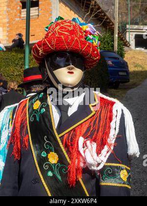 Bagolino Karneval Balari Porträt, Gesicht bedeckt mit der ausdruckslosen elfenbeinfarbenen und schwarzen Maske, Kopf unter einem Filzhut, bedeckt mit rotem Band, Goldschmuck und bunten Bändern, trägt ein schwarzes Kleid mit einem Seidenband mit Blumenstickerei Stockfoto