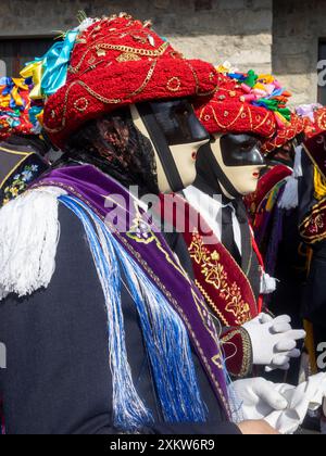 Bagolino Carnival Balari Porträt, Gesicht bedeckt mit der ausdruckslosen elfenbeinfarbenen und schwarzen Maske, Kopf unter einem Filzhut, bedeckt mit rotem Band, Goldschmuck und bunten Bändern, trägt ein schwarzes Kleid mit einem Seidenband mit Blumenstickerei und weißen Handschuhen Stockfoto