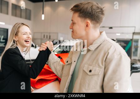 Fröhliche, blonde Managerin im Anzug gratuliert dem männlichen Käufer zum Kauf im Autohaus und gibt dem Kunden Autoschlüssel. Verkäuferin und männlich Stockfoto