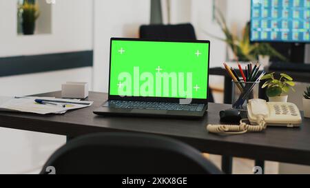 Büro mit grünem Laptop auf dem Schreibtisch und Monitor im Hintergrund mit Börsennotierungen. Mockup-Notebook im Arbeitsbereich und Computerbildschirm im Hintergrund mit Handelsplattform Stockfoto