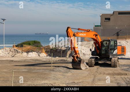 Hitachi Raupenbagger auf einer Baustelle mit dem Atlantik und einem Containerschiff im Hintergrund. Stockfoto