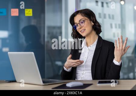 Geschäftsfrau in Anzug, die an Online-Konferenzgesprächen teilnimmt und Ideen mit Begeisterung kommuniziert. Moderne Büroumgebung mit Laptop, Tablet und Haftnotizen im Hintergrund, die eine aktive Arbeitssitzung anzeigen Stockfoto