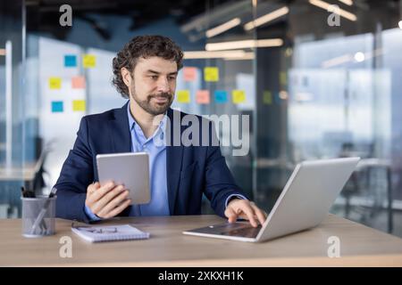 Geschäftsmann in formeller Kleidung, der am Laptop arbeitet und im modernen Büro ein Tablet hält. Arbeitsplatz mit Haftnotizen und Glaswänden. Professionelles Umfeld mit fokussiertem Multitasking für Mitarbeiter Stockfoto