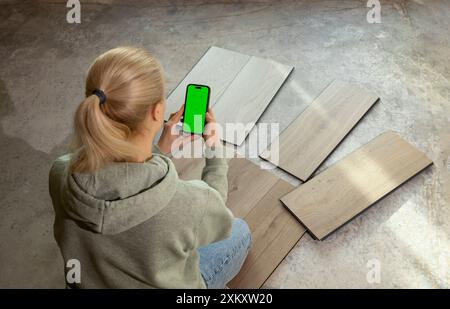 Innenarchitektin hält das Smartphone mit grünem Chroma-Bildschirm über luxuriösen Vinyl-Holzproben auf Betonboden im Haus. Auswählen, Auswählen Stockfoto