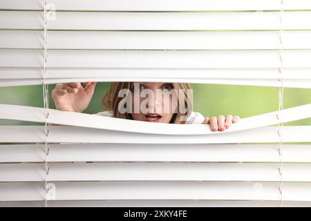 Junge Frau, die durch Fensterläden auf verschwommenem Hintergrund schaut Stockfoto