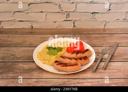 Kartoffelpüree mit gegrillten Würstchen und Tomaten auf einem Holztisch, Besteck in der Nähe. Nahaufnahme. Stockfoto