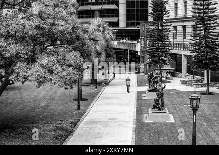 Ein Blick vom Epitaph auf den Betonsteg, während sich ein Stadtbürger seinen Weg durch die Straße macht, und vorbei an Bäumen und Bronzestatuen am ANZAC Platz. Stockfoto