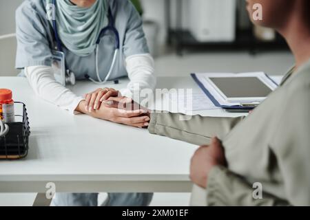 Abgeschnittener Schuss einer jungen Ärztin, die Hand eines schwangeren afroamerikanischen Patienten hält, während beide vor einander sitzen Stockfoto