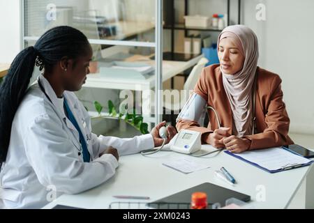Junge afroamerikanische Hausärztin im Labormantel zur Blutdruckmessung muslimischer Frauen in Hijab und Freizeitkleidung Stockfoto
