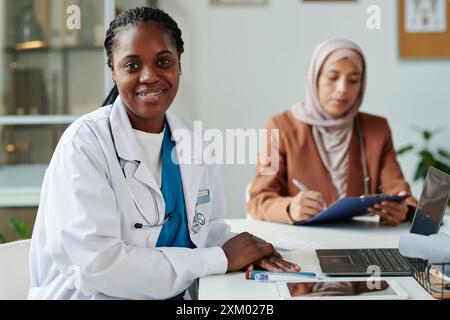 Glückliche junge afroamerikanische Ärztin im Laborkittel, die mit einem Lächeln in die Kamera blickt, während sie am Arbeitsplatz mit Laptop sitzt Stockfoto