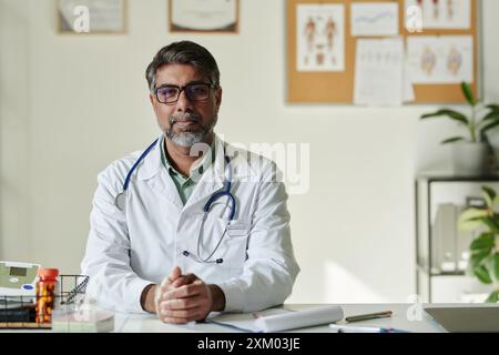 Ernster reifer männlicher Kliniker mit Stethoskop, der in die Kamera schaut, während er am Arbeitsplatz mit medizinischen Dokumenten und anderen Dingen sitzt Stockfoto