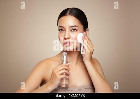 Die lächelnde Frau hält eine Flasche mit kosmetischem Tonikum. Foto einer attraktiven Frau mit perfektem Make-up auf beigefarbenem Hintergrund. Schönheits- und Pflegekonzept. Entfernen Stockfoto