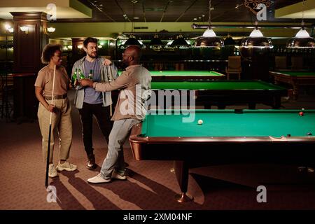 Drei junge interkulturelle Freunde, die mit Flaschen Bier getoast und sich gegenseitig anschauen, während sie am Billardtisch im Spielzimmer stehen Stockfoto