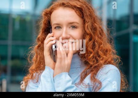 Eine junge Rothaarige genießt eine Frau in weißem Hemd, die mit ihrem Handy spricht und benutzt, in einer modernen Stadt. Lifestyle-Fotos Stockfoto