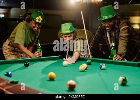 Drei glückliche junge Freunde in grünen Hüten trinken Bier und genießen es, sich zu treffen, während interkulturelle Jungs mit Hinweisen Billard spielen Stockfoto