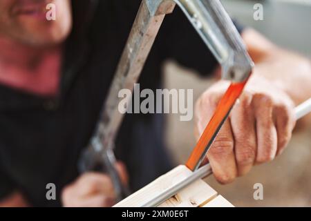 Geschulter Zimmermann, der Metallstäbe mit Bügelsäge in der Werkstatt schneidet und dabei Präzision und Handwerkskunst zeigt. Sägemehl und Holzdielen umgeben ihn als Dämon Stockfoto