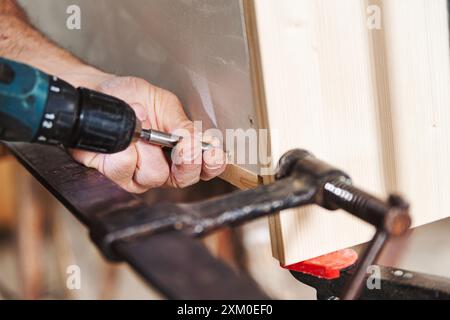 Geschulter Zimmermann mit Akku-Bohrmaschine zum Befestigen von Holzstücken. Präzision und Handwerkskunst in Aktion, Hände konzentriert und stabil. Holzmaserung und Sägedus Stockfoto