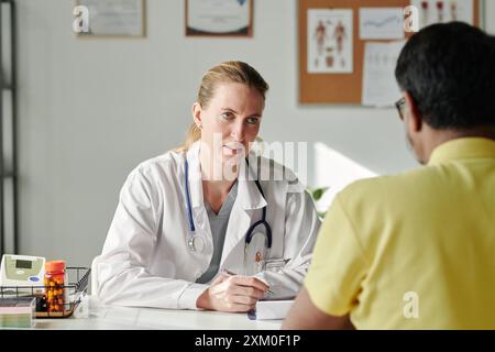 Junge Hausärztin im Laborkittel, die vor ihr sitzenden männlichen Patienten ansieht und ihm ärztlichen Rat gibt Stockfoto