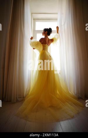 Die Silhouette einer Frau in einem goldenen, luxuriösen Kleid vor dem Hintergrund eines Fensters hält einen Vorhang mit den Händen. Elegante Dame in gelber Seide Stockfoto