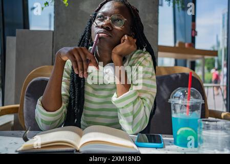 Nachdenkliche afroamerikanische Frau mit Gläsern und Dreadlocks, die im Café sitzt, Notizen in Notizbücher schreibt und Blaugetränk mit Limette genießt. Stockfoto