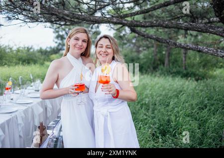 Zwei junge Frauen umarmen und halten Gläser mit Orangencocktail in der Natur. Party im Freien. Charmante Freunde in weißen Kleidern trinken aperol und feiern Stockfoto