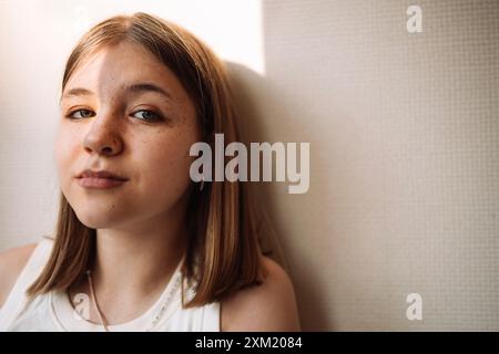 Ein Nahporträt eines süßen Teenagers an der Wand. Ein charmantes lächelndes Mädchen mit Sonnenlicht im Gesicht. Ein hübscher weiblicher Teenager in legeren Klamotten Stockfoto