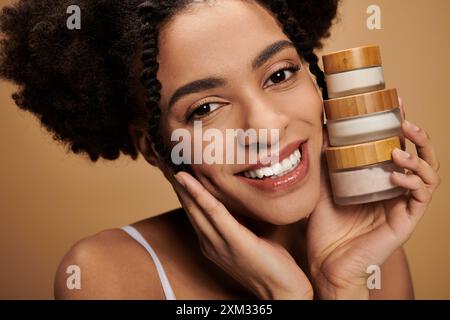 Eine junge afroamerikanische Frau mit einem strahlenden Lächeln hält Hautpflegeprodukte gegen ihr Gesicht. Stockfoto