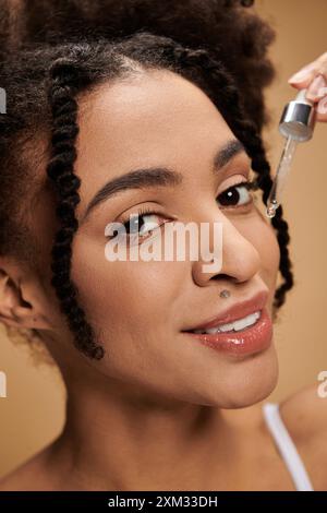 Eine junge afroamerikanische Frau mit schönen braunen Augen und einem hellen Lächeln trägt ein Serum auf ihr Gesicht auf einem weichen beigen Hintergrund. Stockfoto