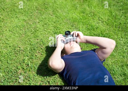 Der Junge liegt auf grünem Gras und macht Aufnahmen mit einer alten und alten Fotokamera. Im Freien. Kopierbereich. Stockfoto