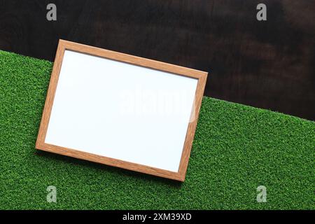 Leerer Rahmen mit weißem Blatt auf dunklem hölzernem Hintergrund mit grünem Kunstrasen. Einfacher Holzrahmen. Platz für Text. Leeres Foto mit Kopierraum. Stockfoto