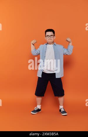 Ein kleiner Junge mit Down-Syndrom beugt seine Muskeln vor einem orangefarbenen Hintergrund. Stockfoto