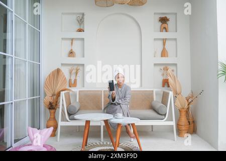 Glückliche asiatische Frau, die einen grauen Bademantel trägt, auf einer Couch sitzt und den Bildschirm des Smartphones zeigt, während sie fröhlich lächelt. Urlaubsangebot Konzept. Stockfoto