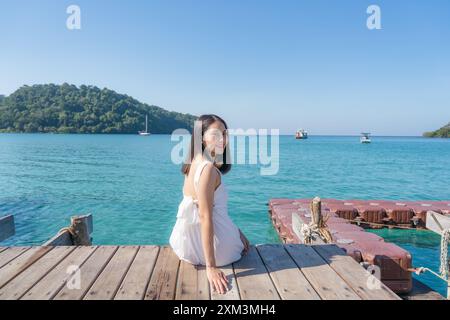 Schöne junge asiatische Frau in weißem Kleid, die auf hölzernem Pier zwischen tropischem Meer im Sommerurlaub auf Koh Kood Island sitzt Stockfoto