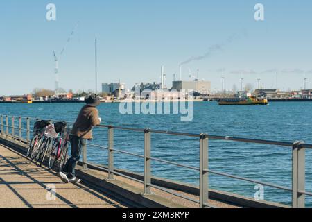 Einsamer männlicher Tourist, der an sonnigen Tagen am Wasser mit Industrieanlagen und Windturbinen genießt und sich entspannt Stockfoto