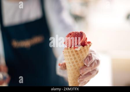 Waffelkegel mit köstlichem Erdbeereis in der Hand. Der Verkäufer gibt eine Eiskugel in eine Waffelkugel für Löffel Stockfoto