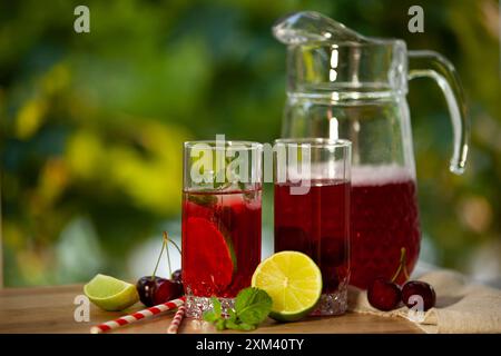 Ein Glaskrug mit erfrischendem Fruchtpunsch, begleitet von zwei Gläsern, garniert mit Limette, Minze und Kirschen. Horizontales Foto. Stockfoto