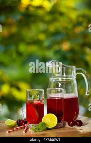 Ein Glaskrug mit erfrischendem Fruchtpunsch, begleitet von zwei Gläsern, garniert mit Limette, Minze und Kirschen. Vertikales Foto. Stockfoto