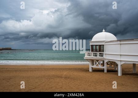 Das ikonische Badehaus Balneario de La Palma am Strand Playa de La Caleta in Cadiz, Andalusien, Spanien, unter einem dramatisch bewölkten Himmel. Stockfoto