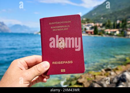 Malcesine, Gardasee, Italien - 20. Juli 2024: Hand hält im Urlaub deutscher Reisepass vor den Gardasee in Italien *** Hand hält im Urlaub deutscher Reisepass vor den Gardasee in Italien Stockfoto