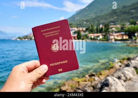 Malcesine, Gardasee, Italien - 20. Juli 2024: Hand hält im Urlaub deutscher Reisepass vor den Gardasee in Italien *** Hand hält im Urlaub deutscher Reisepass vor den Gardasee in Italien Stockfoto