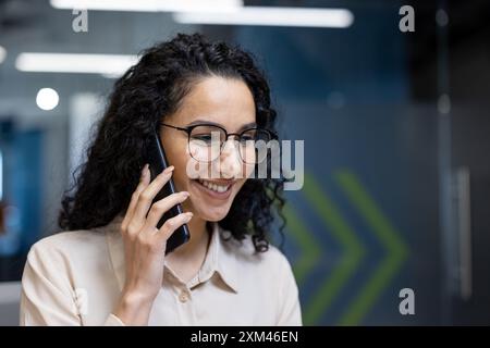 Lächelnde Geschäftsfrau mit Brille und lockigem Haar, die im modernen Büro auf dem Smartphone spricht. Professionelles Personal, das sich in freundlichen Gesprächen engagiert und Kommunikation und Führung am Arbeitsplatz repräsentiert. Stockfoto