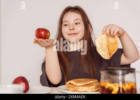 Ein kleines Mädchen hält einen Pfannkuchen in der einen Hand und einen roten Apfel in der anderen und schaut ihn an. Konzept der Wahl des richtigen Essens. Hochwertige Fotos Stockfoto